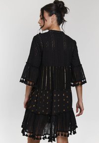 Abito di pizzo nero con design a strati, caratterizzato da maniche a campana, dettagli dorati e rifiniture con frange, che mette in mostra motivi ad intaglio.