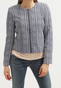 Granatowo-biała kurtka w geometryczny wzór o skróconym kroju, z okrągłym dekoltem, długimi rękawami i bocznymi kieszeniami; wzór geometryczny na teksturowanej tkaninie.