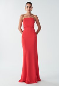 Punainen olkaimeton iltapuku, jossa on tiukka vartalonmyötäinen yläosa, virtaava hame ja sileä pinta. Korostaa hyvin määriteltyä vyötärölinjaa ja on lattiaan asti ulottuva.