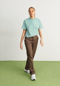 T-shirt en coton vert clair avec un petit logo, associé à un pantalon marron et des baskets blanches. Le modèle se tient sur un sol vert.