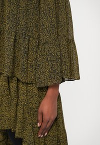 Černá a hořčicově žlutá látka s paisley vzorem a volány; viditelná ruka jemně drží materiál. Měkká textura, lehký design.