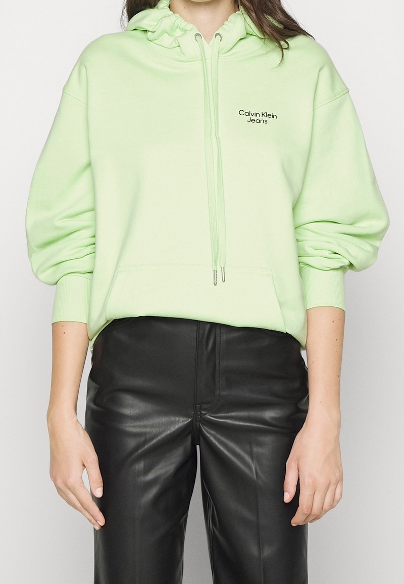 Persoon die een lichtgroene Calvin Klein hoodie draagt met een voorvak en zwarte nepleren broek, staand met ontspannen armen.
