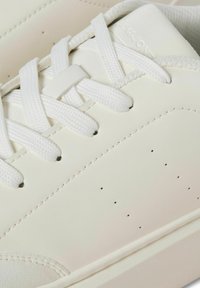 White leather sneaker with textured surface, rounded toe, and white laces. Features small ventilation holes and discreet branding on the side.
