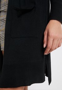 Svart stickad cardigan med sidoslits, ribbade ärmslut och en stor framficka på vänster sida. Texturerad tyg med en slät yta.