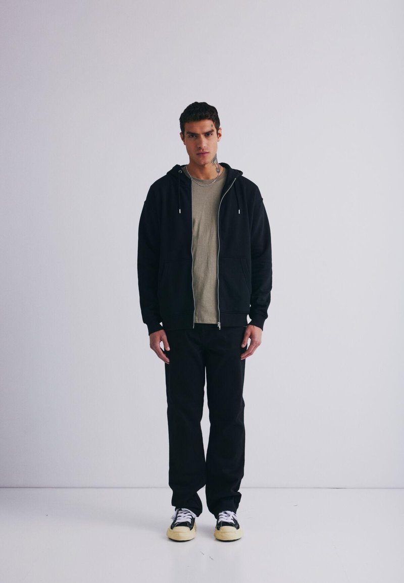 Felpa nera con zip sopra una maglietta grigia chiara, abbinata a pantaloni neri e sneakers nere con suole gialle. Design semplice, dettagli minimi.