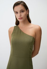 Asymmetrisches Ein-Schulter-Kleid in Olivgrün, aus strukturiertem Stoff; enganliegendes Oberteil, das in einen ausgestellten Rock übergeht. Goldene Ohringe.