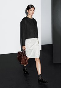 Kvinna går i svart knäppt jacka, vit knälång kjol, svarta oxfordskor, genomskinliga strumpor, bär en stor brun läderhandväska.