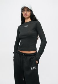 Femme aux longs cheveux foncés portant une casquette blanche, un crop top noir à manches longues côtelées et un large pantalon de jogging noir avec des logos "Jordan".