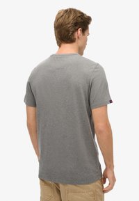 Camiseta gris de manga corta con cuello redondo. Tejido de algodón suave. Presenta un pequeño parche con el logo en la manga izquierda. Diseño simple y sin adornos.