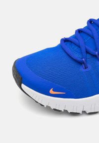 Plava tekstilna atletska cipela s crnim đonom i bijelim srednjim đonom, s ljubičastim vezicama i narančastim Nike logotipom sa strane.