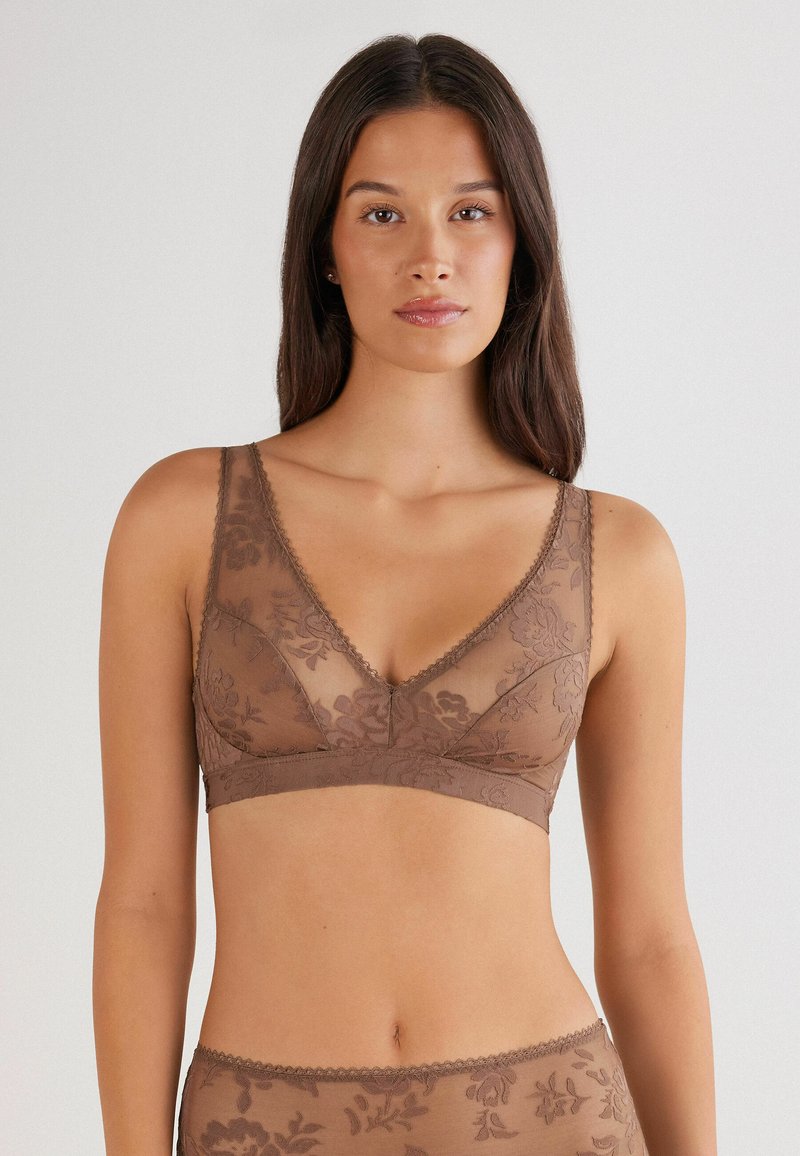 Femme portant un bralette en dentelle florale marron et un bas assorti sur un fond clair uni, se tenant de face avec une expression neutre.