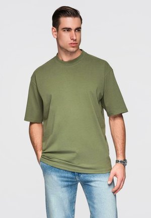 Giovane uomo con una T-shirt oversize verde oliva e jeans azzurri chiaro, in piedi con le mani parzialmente nelle tasche, che guarda di lato.