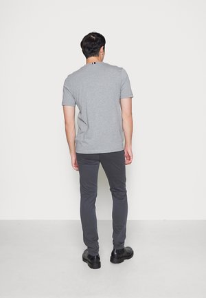 Mann steht mit dem Rücken zur Kamera, trägt ein graues kurzärmeliges Hemd, dunkelgraue Hose und schwarze Schuhe vor einem einfarbigen Hintergrund.