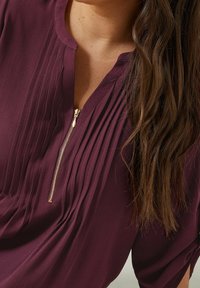 Gros plan sur une femme portant un blouse de couleur prune avec des plis verticaux et une fermeture éclair dorée au niveau du col, avec de longs cheveux bruns tombant sur une épaule.