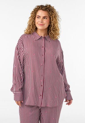 Mujer de cabello rizado que viste una camisa y pantalones a juego, de gran tamaño, con rayas verticales en burdeos y blanco, con pequeños puntos bordados en rosa.