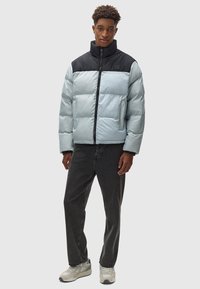 Daunenjacke in Hellblau mit schwarzem Kragen und Frontreißverschluss, ausgestattet mit gesteppten Abschnitten und Seitentaschen. Getragen mit dunklen Jeans und Sneakern.