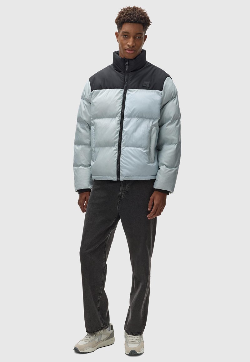 Daunenjacke in Hellblau mit schwarzem Kragen und Frontreißverschluss, ausgestattet mit gesteppten Abschnitten und Seitentaschen. Getragen mit dunklen Jeans und Sneakern.