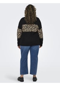 Črn pulover z leopardjim vzorcem na spodnjem delu, dolgi rokavi in sproščen fit. V kombinaciji z modrimi hlačami z ravnimi nogavicami in črnimi čevlji.