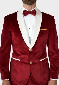 Ein roter Samt-Smokingjackett mit weißen Satin-Revers, drei goldenen Knöpfen und einem gelben Taschentuch. Verfügt über einen strukturierten Stoff und seitliche Taschen.