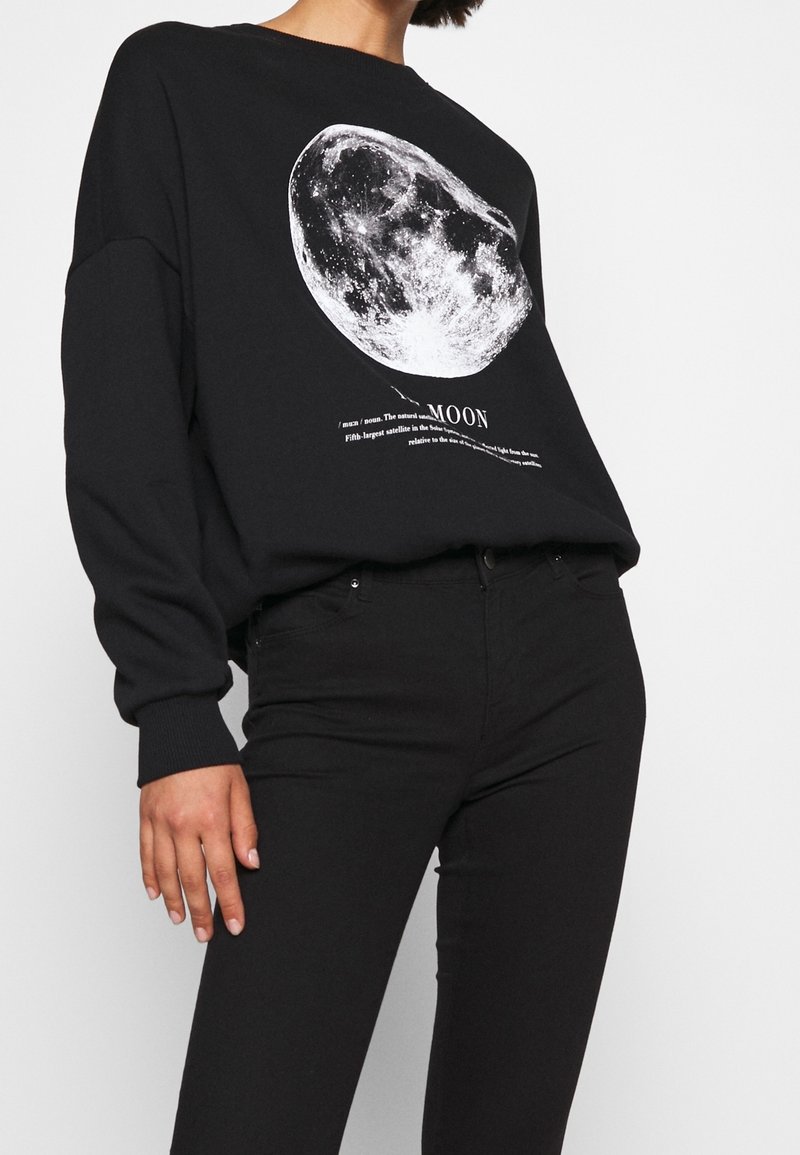 Personne portant un sweat-shirt noir avec un graphisme détaillé de la lune blanche et un texte, associé à un pantalon noir sur un fond uni.