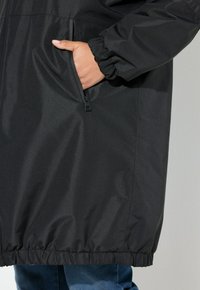 Hand in zijzak van zwarte ritsjacket met elastische manchetten, gedragen over blauwe spijkerbroek.