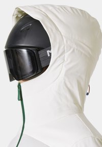 Witte skijas met een capuchon, voorzien van een groene rits en een gestructureerde stof. Een zwarte helm wordt gedragen, uitgerust met donkere skibril.