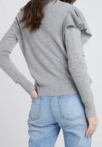 Suéter de punto gris claro con detalles de volantes en los hombros, mangas largas y dobladillo acanalado. Combinado con jeans de mezclilla azul claro. Diseño minimalista.