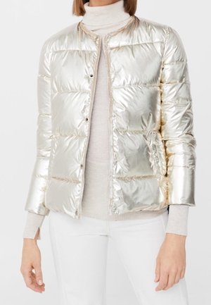 Femme portant une veste matelassée argentée métallique brillante par-dessus un pull à col roulé beige et un pantalon blanc.