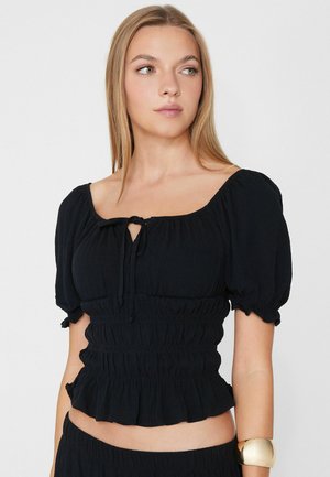 Junge Frau mit langen blonden Haaren trägt ein schwarzes, kurz geschnittenes Top mit Puffärmeln und einer Schleifendetail am Ausschnitt sowie ein goldenes Manschettenarmband.
