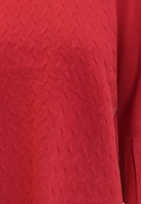 Röd texturerad stickad topp med ett zickzackmönster, långa ärmar och en mjuk yta. Tyget verkar vara lätt och andningsbart.