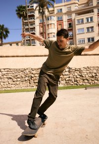 Un skateboard con una tavola in legno scuro. La persona indossa una maglietta verde oliva e pantaloni scuri, in equilibrio sullo skateboard all'aperto.