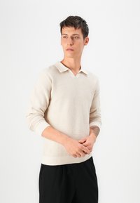 Beige strikkegenser med V-hals og kontrasterende krage; lange ermer med ribbestrikkede mansjetter; brukt med sorte bukser.