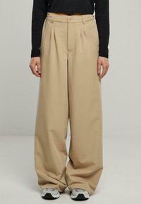 Pantalon large beige avec plis avant, tissu texturé et bouton à la taille. Associé à des baskets grises sur un fond uni.