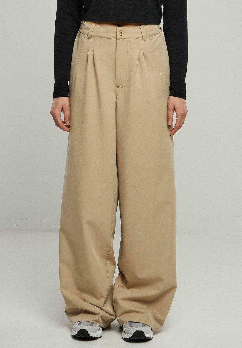 Pantalon large beige avec plis avant, tissu texturé et bouton à la taille. Associé à des baskets grises sur un fond uni.