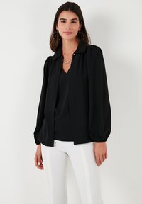 Black blouse with balloon sleeves, a collared neckline, and a button detail. The material appears smooth and lightweight. Worn over a black top.