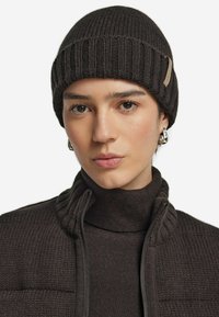 Mörkbrun stickad mössa med ribbat mönster, som har en liten etikett på sidan och bärs tillsammans med en matchande polotröja.