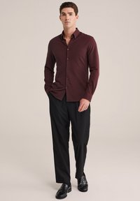 Burgundy színű, hosszú ujjú ing gombokkal, fekete nadrággal és fekete cipővel párosítva. Az ingnek van gallérja és karcsú szabása.