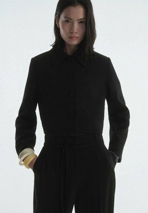 Veste noire courte avec un large col, fermeture par boutons et manches longues. Associée à un pantalon noir taille haute et des bracelets en bangles superposés.