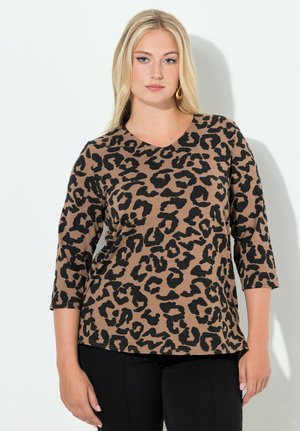 Kobieta o długich blond włosach, ubrana w brązową bluzkę w panterkę z rękawami do łokcia oraz czarne spodnie, stojąca na białym tle.