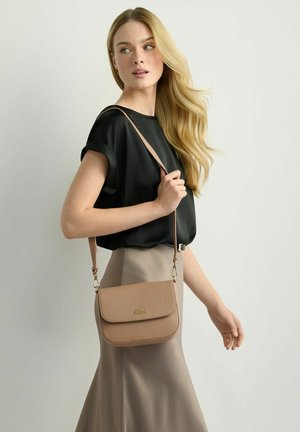 Kobieta o długich blond włosach, ubrana w czarną bluzkę i beżową spódnicę, nosząca na ramieniu rdzawobrązową skórzaną torebkę na ramię.