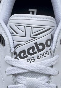 White leather sneaker with flat white laces, featuring a black "Reebok" logo and "BB 4000 II" text on a padded tongue with a Union Jack design.