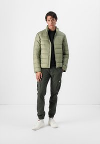 Olijfgroene pufferjack met gecapitonneerde textuur, ritssluiting en hoge kraag, gecombineerd met donkere cargobroek en witte sneakers.