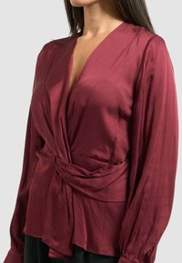 Blusa di seta borgogna con maniche lunghe, scollatura a V profonda e dettaglio frontale arrotolato che crea un effetto arricciato in vita.
