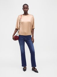 Femme aux cheveux courts portant un top plissé beige, un jean bleu, des ballerines noires pointues, et tenant un petit sac rond rouge avec une bandoulière chaîne.
