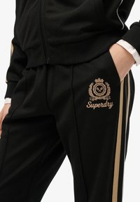 Sort træningsdragt med gulddetaljer. Har et broderet logo og striber langs siderne. Materialet har en glat tekstur, designet til komfort.