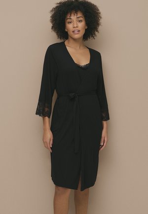 Robe noire en tissu doux, avec des manches bordées de dentelle et une ceinture à nouer. Présentée sur un fond neutre.