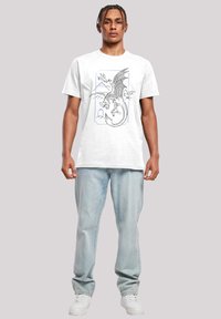 Camiseta de algodón blanca con un gráfico de dragón negro y azul con un fondo montañoso. Combinada con jeans de mezclilla azul claro y zapatillas blancas.