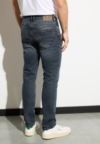 Mężczyzna w ciemnoszarych dżinsach i białych trampkach, stojący w pomieszczeniu przy białej ścianie na betonowej podłodze.