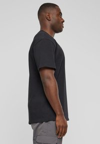 T-shirt en coton noir à manches courtes, col rond et coupe décontractée, porté par une personne faisant face à gauche. Détail de logo flou près du col.