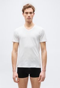 Jeune homme aux cheveux bouclés portant un t-shirt blanc uni à col en V et un boxer noir, debout devant un fond blanc.